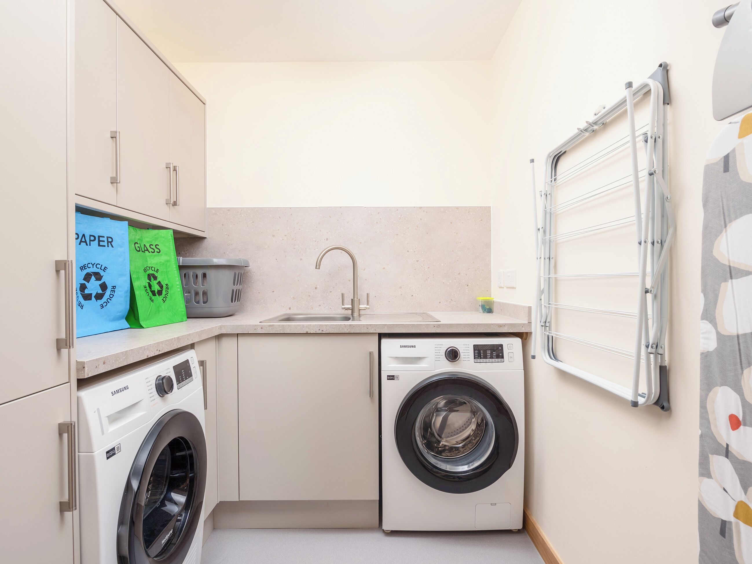 A compact laundry room with a washing machine, a sink, and recycling bags for paper and glass.