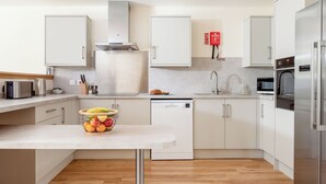 Una cocina moderna con armarios blancos, electrodomésticos de acero inoxidable, una isla central con fruta y un extintor de incendios en la pared.