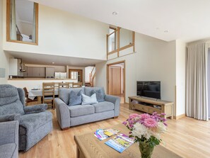 A spacious living area with a grey sofa, a wooden coffee table, and a TV. A dining area is visible in the background with a wooden table and chairs. - Largo Lodge - UKC5801 (Lundin Links)