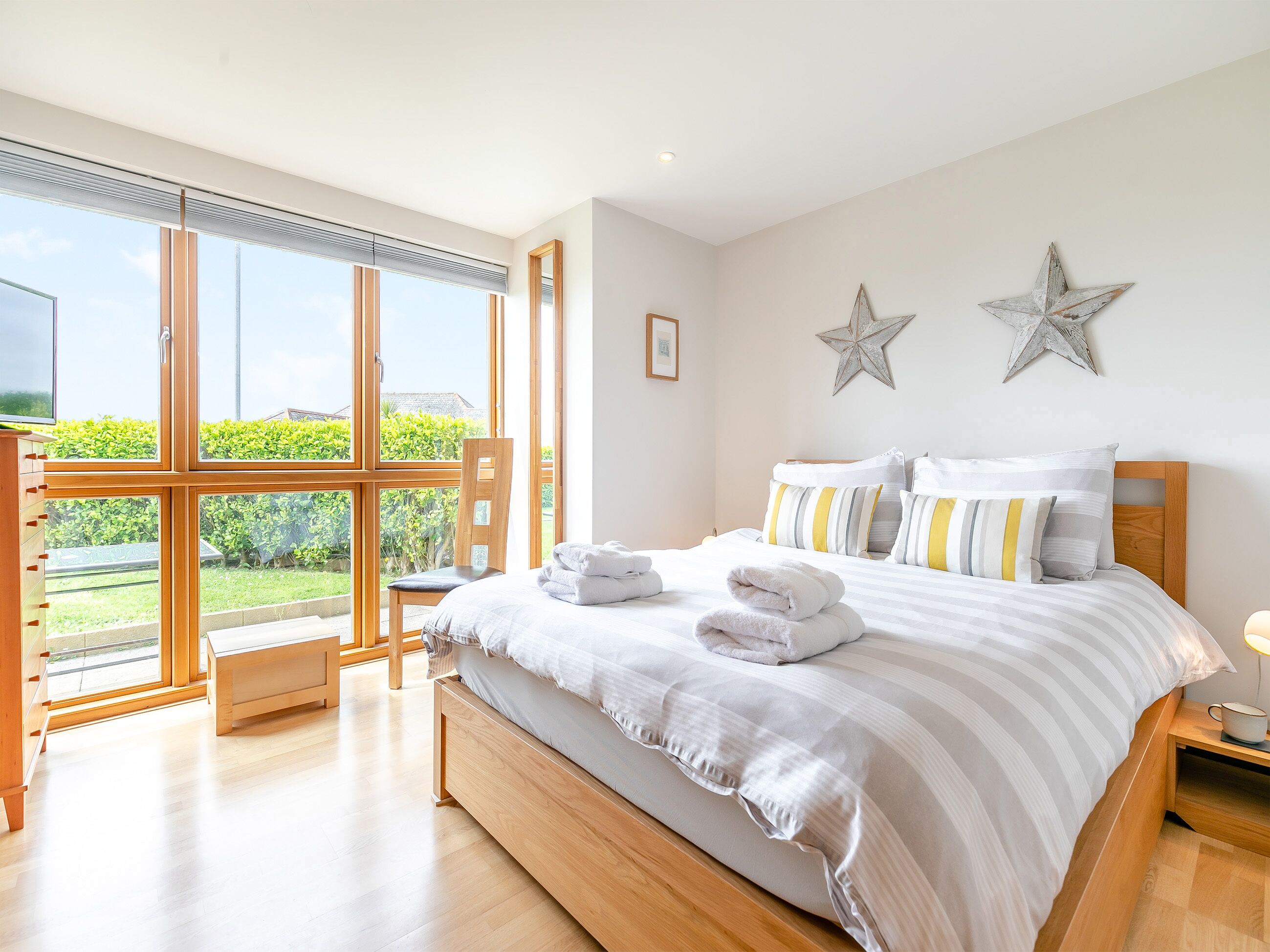 A bedroom with a large bed, a wooden nightstand, a chair, and a view of greenery through large windows.