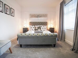 A bedroom with a tufted headboard, a large bed, two bedside tables with lamps, a grey upholstered bench, and a window with curtains.