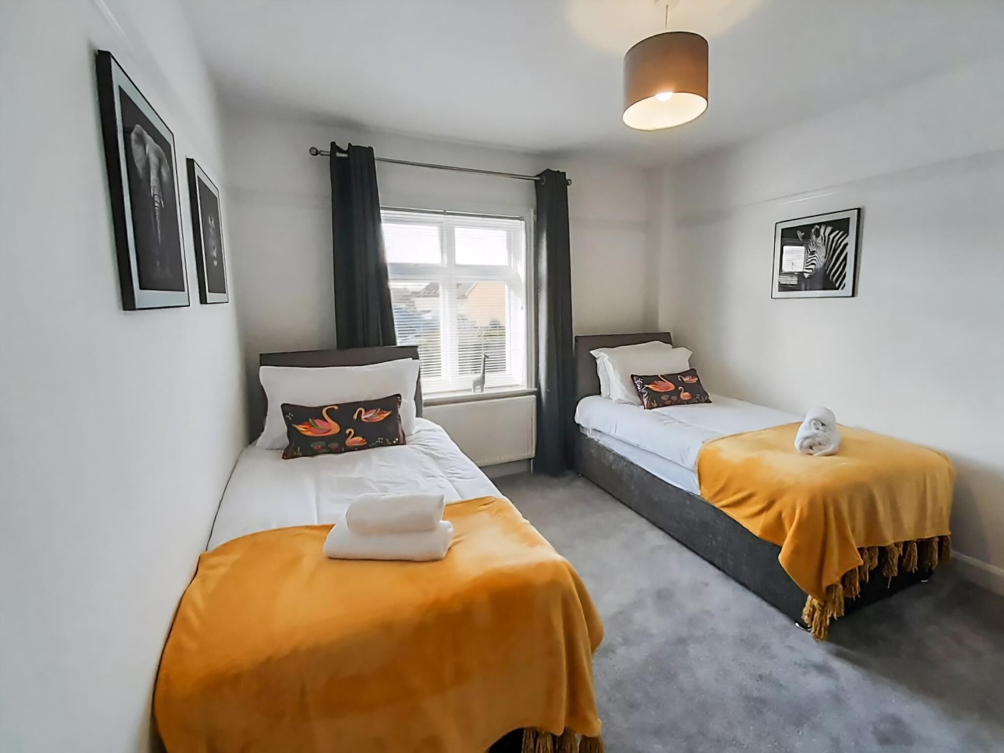 A hotel room with two beds, a window with curtains, a hanging light fixture, and framed pictures on the walls.