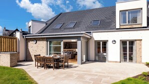 Ein modernes Haus mit einer Terrasse, einem Essbereich und einer durch Glastüren einsehbaren Küche.