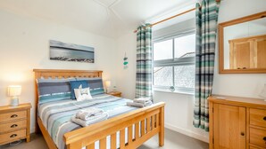 A bedroom with a wooden bed, a nightstand with a lamp, a wooden dresser, and a window with curtains. - Hollyhocks is a charming cottage that is over 100 years old. (St Minver, near Rock)