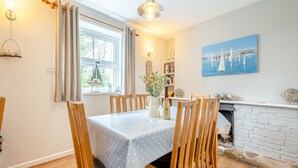 A dining area with a table set for four, wooden chairs, a chandelier, a window with curtains, a bookshelf, and a painting of a harbor. - Hollyhocks is a charming cottage that is over 100 years old. (St Minver, near Rock)
