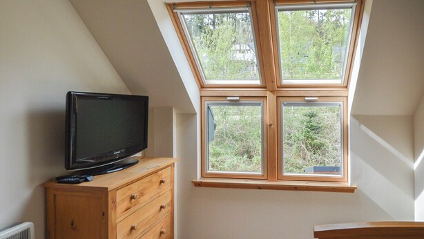 Ein Zimmer mit schrägem Dach, einer hölzernen Kommode mit Fernseher darauf und einem Fenster mit Blick auf Grünflächen.
