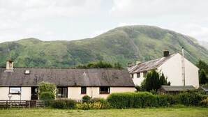Ett hus på landsbygden med ett sluttande tak, omgivet av en välskött trädgård och ett berg i bakgrunden.