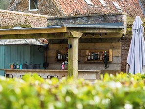 A wooden outdoor bar area with shelves of bottles, a covered seating area, and a brick building in the background. - 7 bedroom accommodation in Sedgeford, near Hunstanton (Sedgeford, near Hunstanton)
