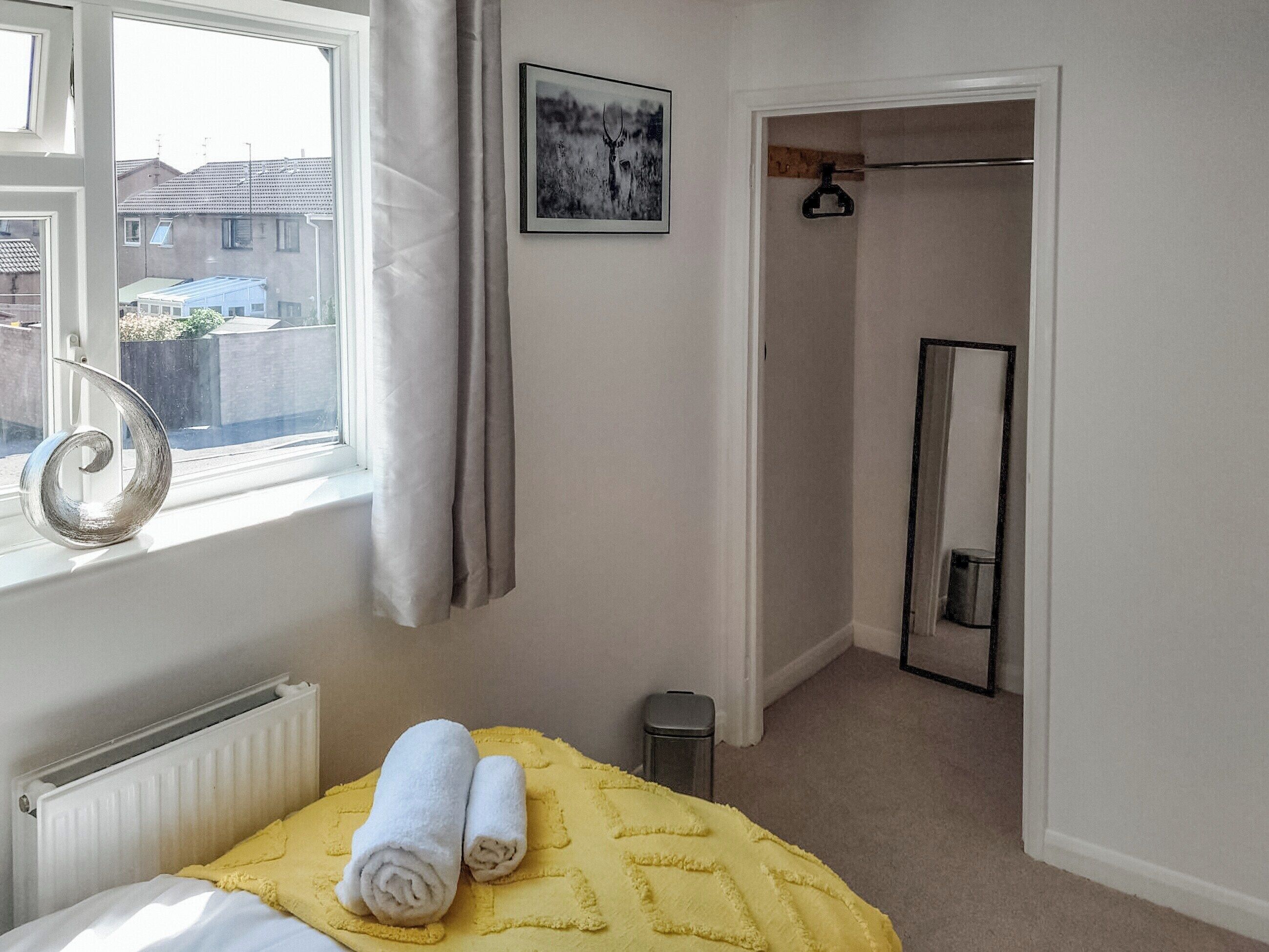 A bedroom with a bed, a window with curtains, a radiator, a yellow bedspread, a towel, a small trash bin, a framed picture on the wall, and a mirror.
