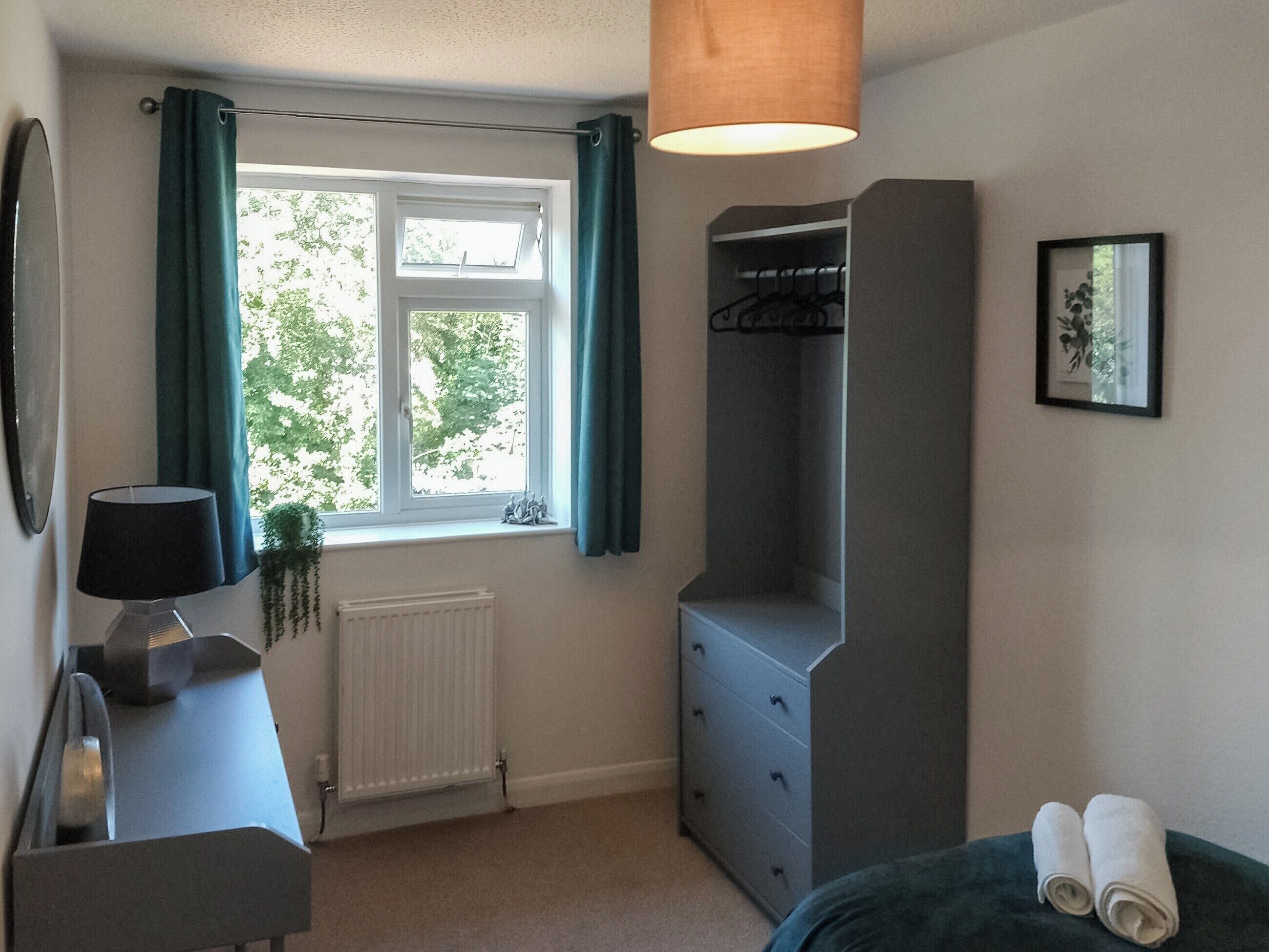 Ein Schlafzimmer mit Fenster, Heizkörper, Schreibtisch mit Lampe, Kleiderschrank und einem Bett mit zusammengerollten Handtüchern.