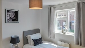 Une chambre à coucher avec un lit, une table de chevet avec une lampe, un radiateur, une fenêtre avec des rideaux et un tableau encadré au mur.