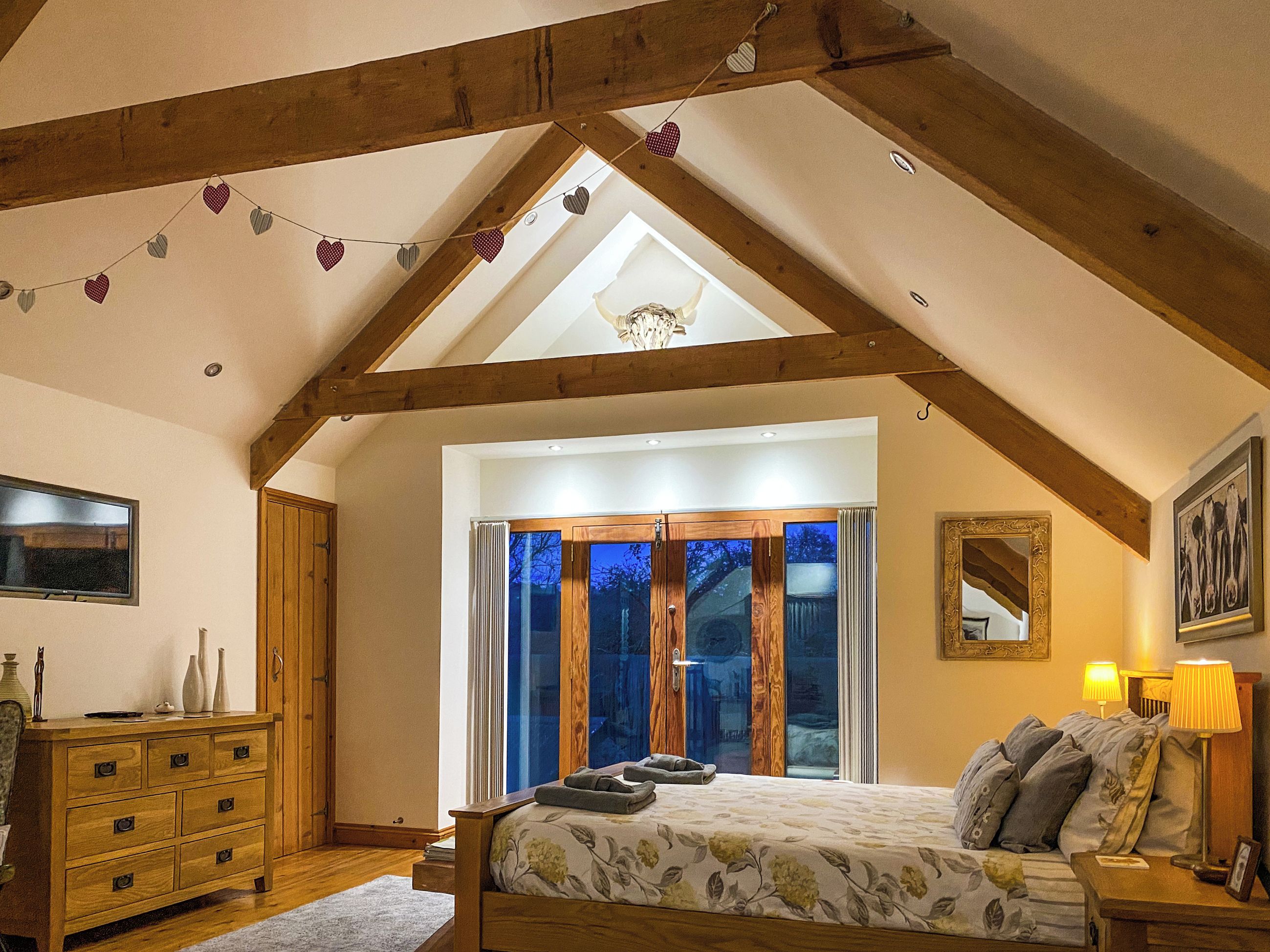 Ein Schlafzimmer mit geneigter Decke, Holzbalken, einem Bett mit Blumenmuster, einem Nachttisch mit Lampen und einer Kommode.