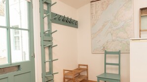 Una stanza con una scala in legno, due sedie di legno, una piccola mensola in legno, una mappa appesa al muro e una porta con un oblò.
