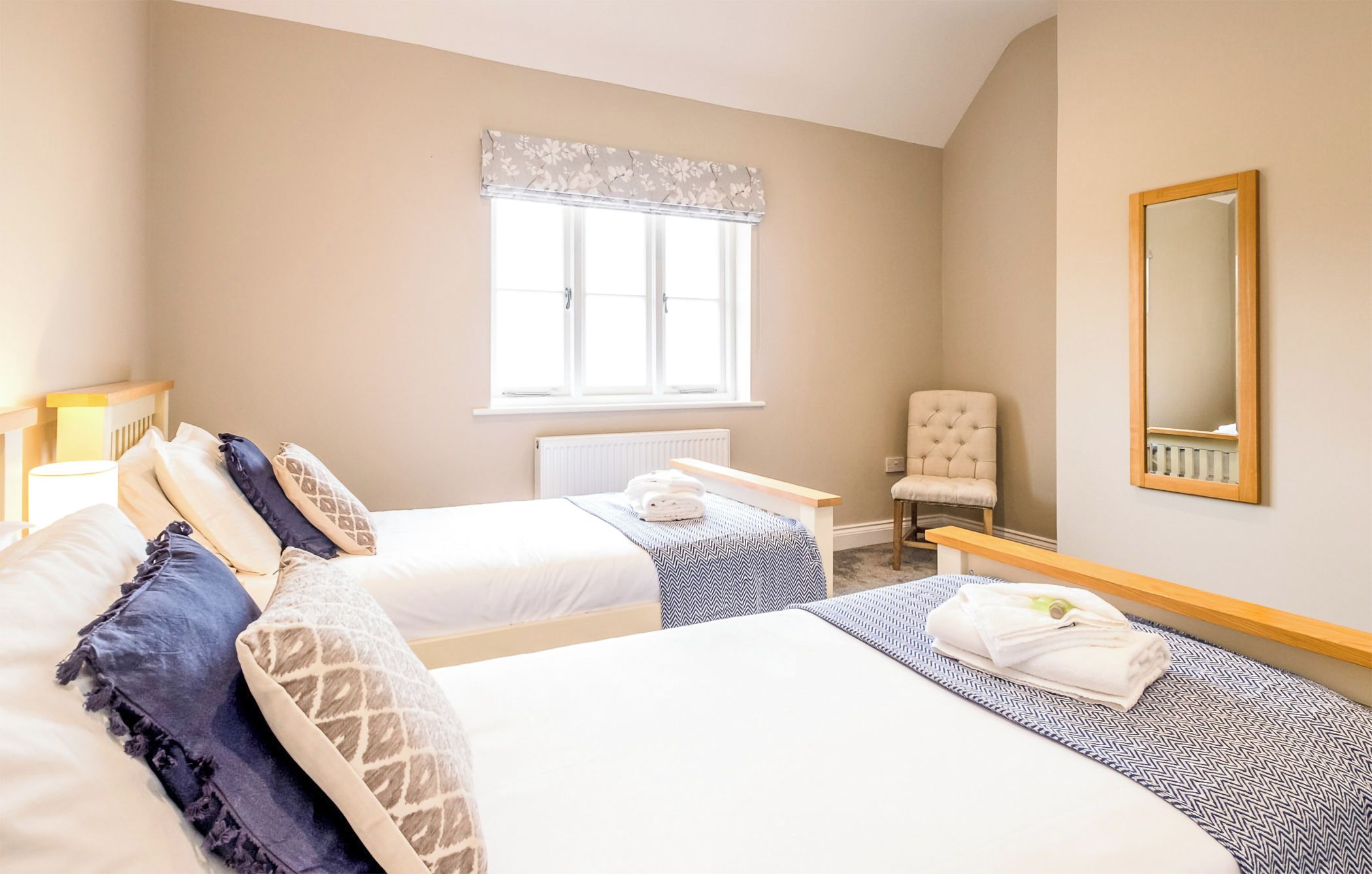A hotel room with two beds, a chair, a window with a floral valance, and a mirror on the wall.