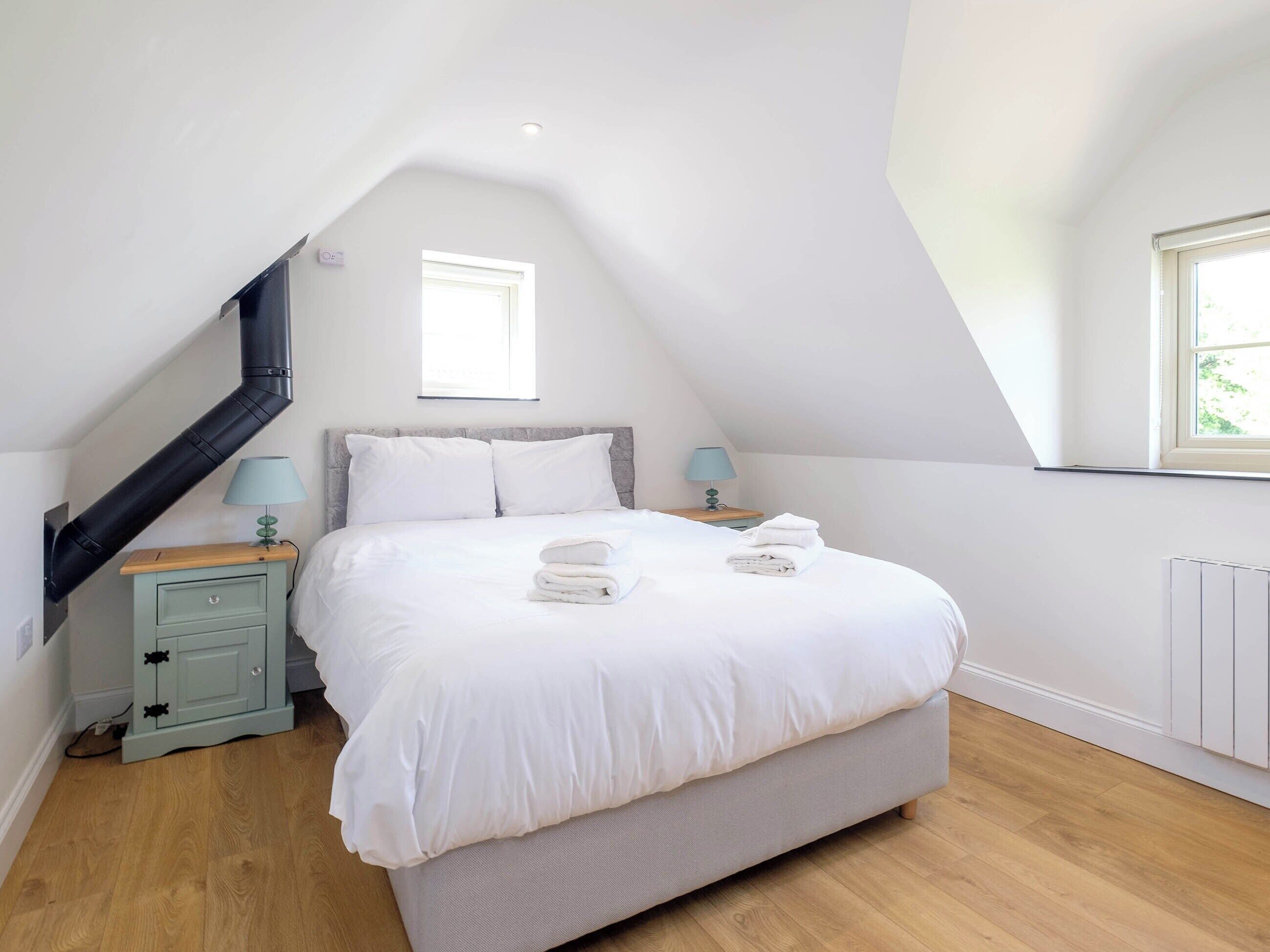 Une chambre avec un plafond en pente, un lit avec une literie blanche, deux tables de chevet avec des lampes et une fenêtre.