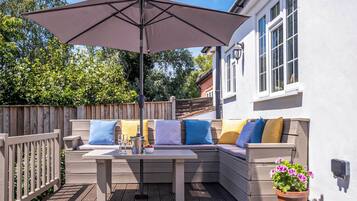 An outdoor patio with a wooden deck, a cushioned bench with colorful cushions, a table with a bottle and glasses, and a potted plant.