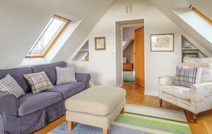 A loft-style living room with a purple sofa, a white armchair, and a small ottoman. There are skylights and a wooden door leading to a bedroom. - White Cottage Apartment (Iken)