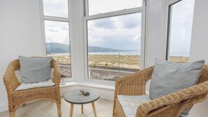 Zwei Korbstühle mit grauen Kissen und ein kleiner runder Tisch stehen vor großen Fenstern mit Blick auf einen Strand und die Ferne.