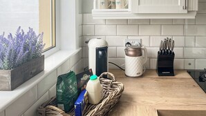 Ett kök med vita skåp, ett fönster med utsikt över hus, en kaffebryggare, en vattenkokare, ett knivställ och en korg med matvaror.