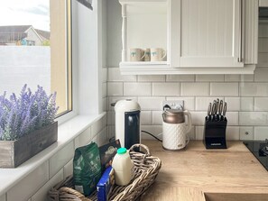 A kitchen with white cabinets, a window with a view of houses, a coffee maker, a kettle, a knife block, and a basket with groceries. - 2 bedroom accommodation in Anstruther (Anstruther)