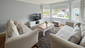 Ett modernt vardagsrum med en platt-TV, två soffor, ett soffbord och stora fönster med utsikt över gatan.
