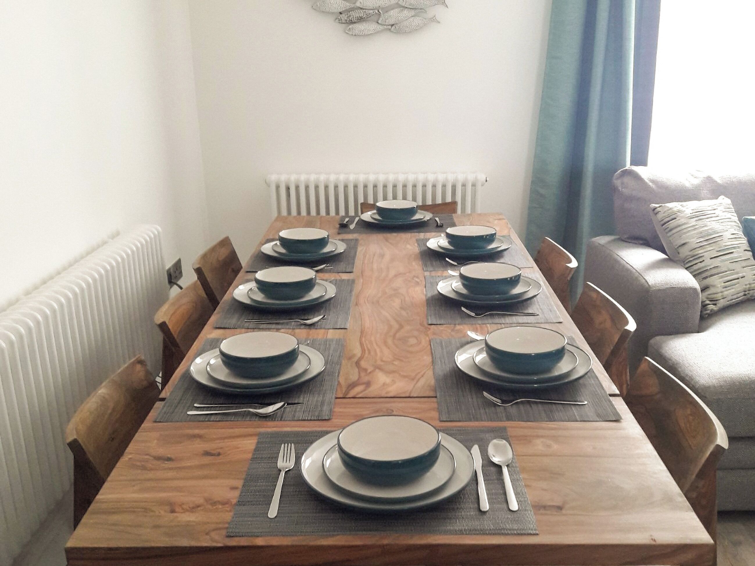 Una mesa de comedor preparada con platos, fuentes, cubiertos y manteles individuales, flanqueada por sillas y un radiador.