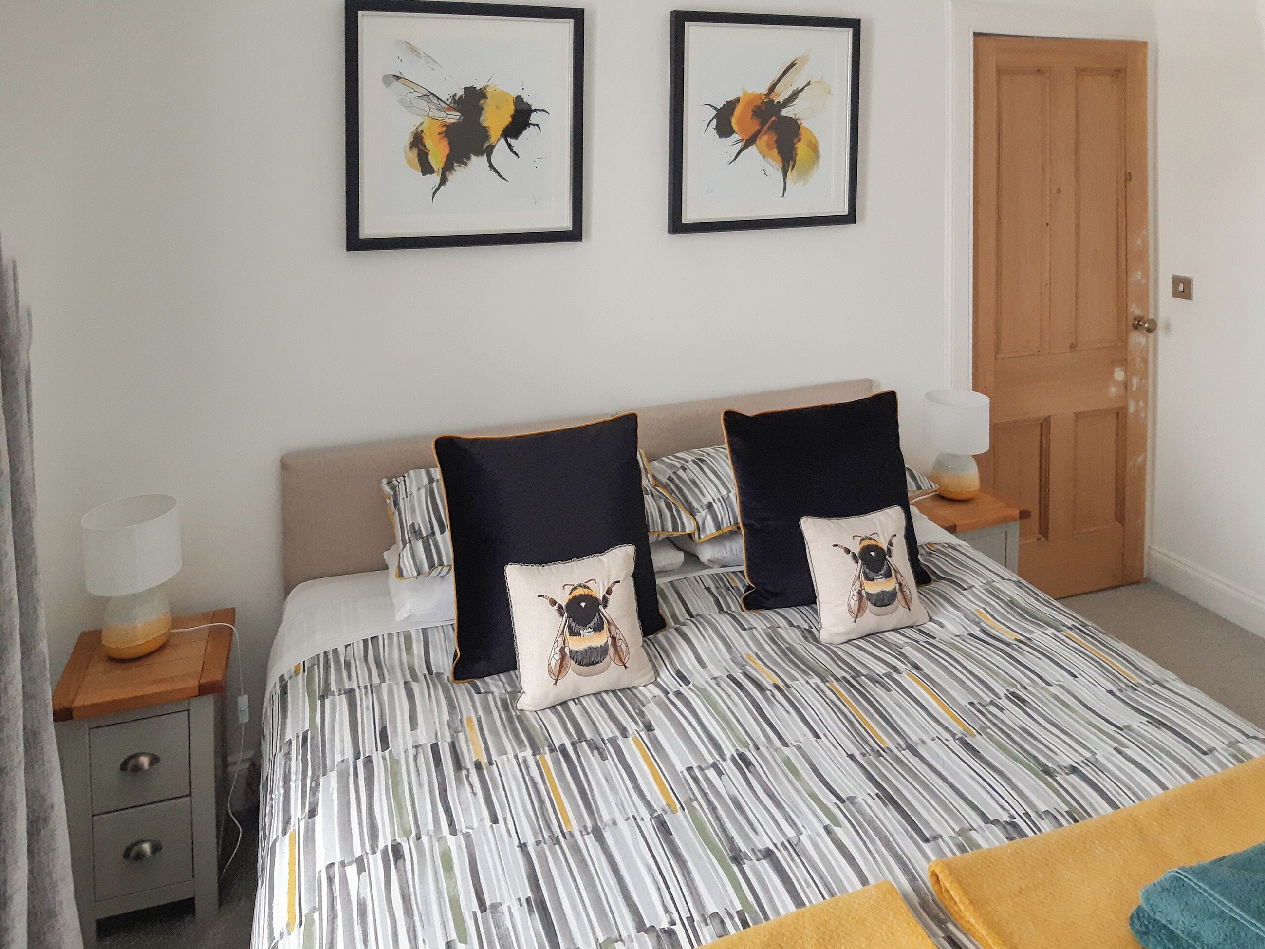 A bedroom with a bed, two pillows with bee designs, a bedside table with a lamp, and a door.