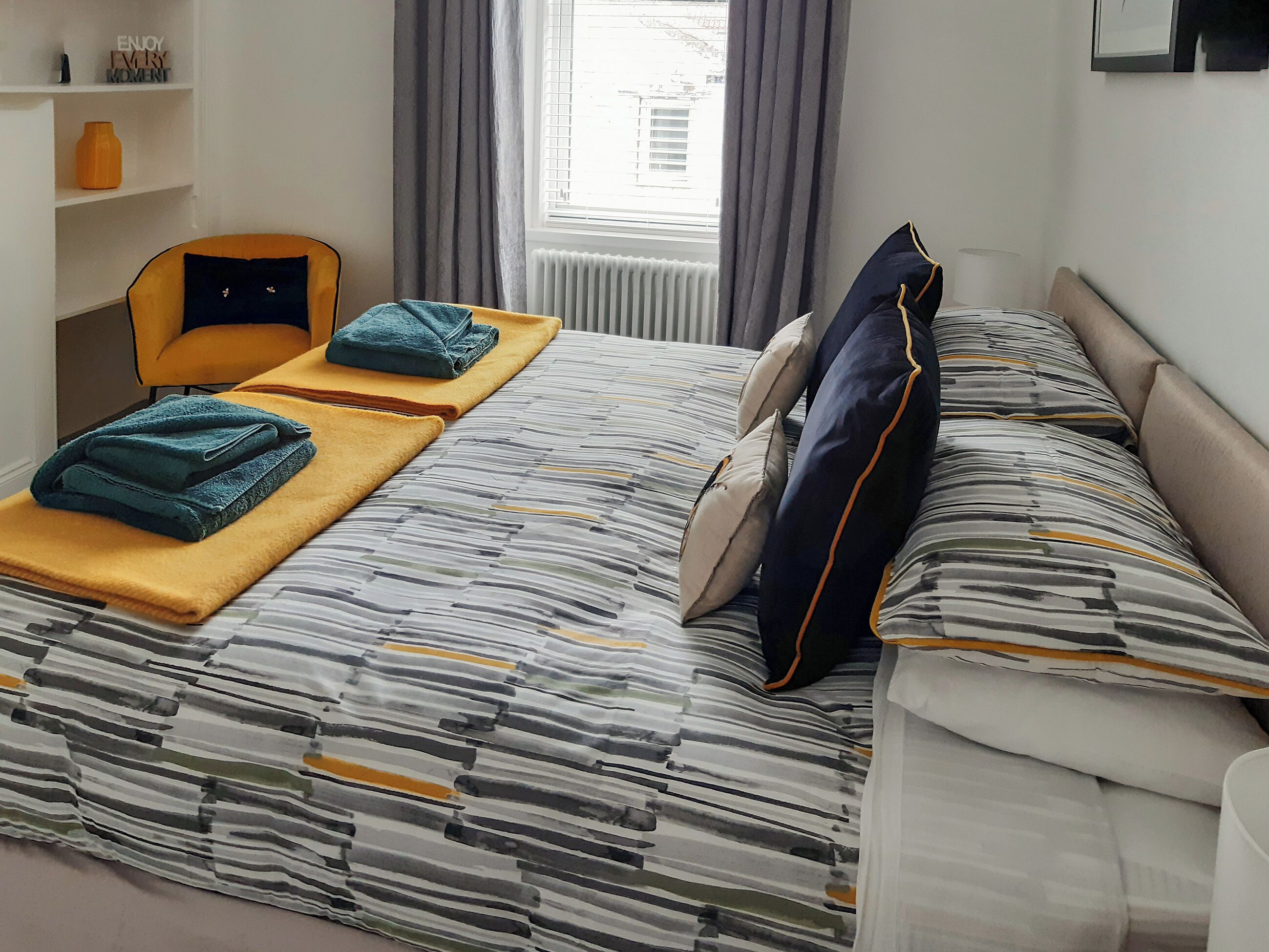 A neatly made bed with striped bedding, a yellow chair, and a small table with folded towels.