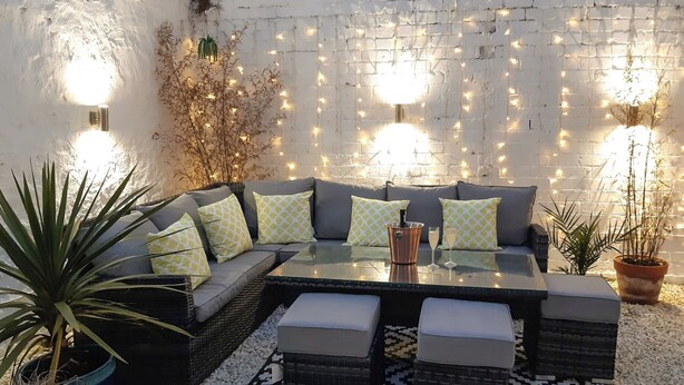 Un espace extérieur accueillant, aménagé avec un canapé et des poufs, une table en verre et des guirlandes lumineuses décoratives, le tout adossé à un mur de briques blanches.