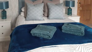 Un lit bien fait, recouvert d’une couverture bleue et agrémenté de deux serviettes pliées, une tête de lit ornée de coussins à motifs et deux lampes de chevet posées sur des tables de nuit blanches.