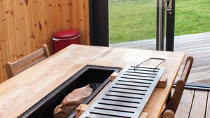 Une table en bois dotée d’un barbecue intégré, une poubelle rouge et une vue sur une zone verdoyante, visible à travers une porte vitrée.