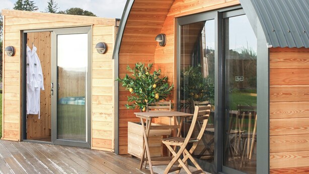 Une cabane en bois avec une porte en verre, une petite table et une chaise à l’extérieur.