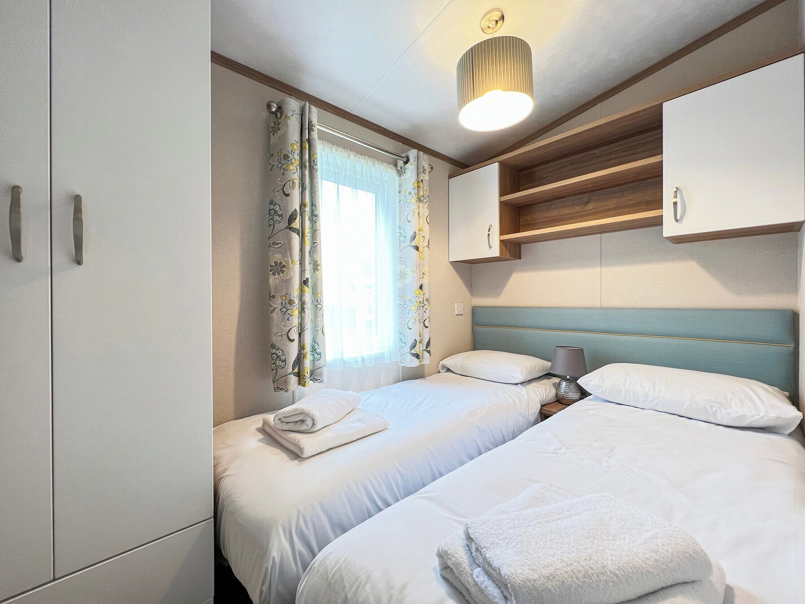 A bedroom with two beds, a window with floral curtains, a bedside lamp, and a ceiling light fixture.
