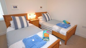 Habitación de hotel con dos camas individuales, cada una con cabecero de madera, ropa de cama blanca y almohadas a rayas. Se observan mesitas de noche con lámparas, un cuadro pequeño en la pared y una toalla azul sobre una de las camas.