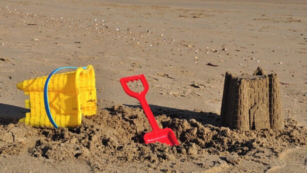 Una playa arenosa con un cubo amarillo para hacer castillos, una pala roja y una estructura de castillo de arena.