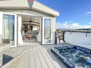 A modern outdoor deck with a hot tub, a seating area with a sofa and table, and a glass door leading to a kitchen and living space. - 2 bedroom accommodation in St Andrews (St Andrews)