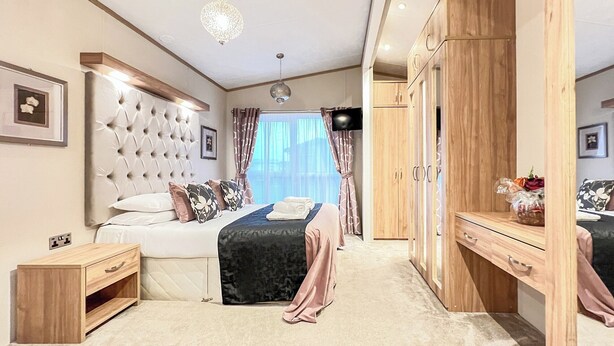 Une chambre à coucher avec une tête de lit capitonnée, un lit avec une tête de lit sombre, une armoire en bois et un téléviseur fixé au mur.