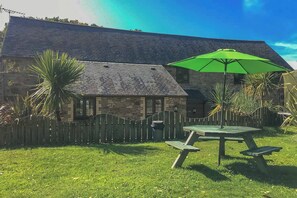 A stone building with a sloped roof, a wooden fence, and a picnic table with a green umbrella. - Fistral Cottage - CWC46839 (Perranporth)