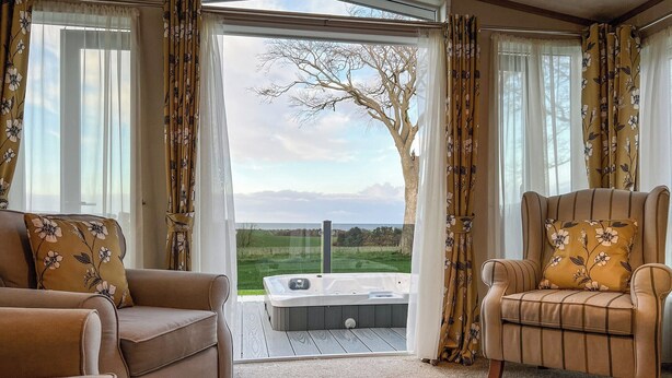 A cozy living room with a sofa, armchair, and a view of a green landscape through an open door. - 2 bedroom accommodation in St Andrews (St Andrews)