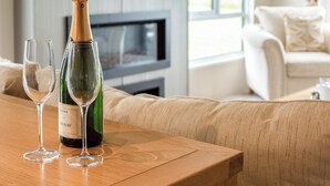 Eine Weinflasche und zwei Gläser auf einem Holztisch in einem modernen Wohnzimmer mit Sofa und Fernseher im Hintergrund.