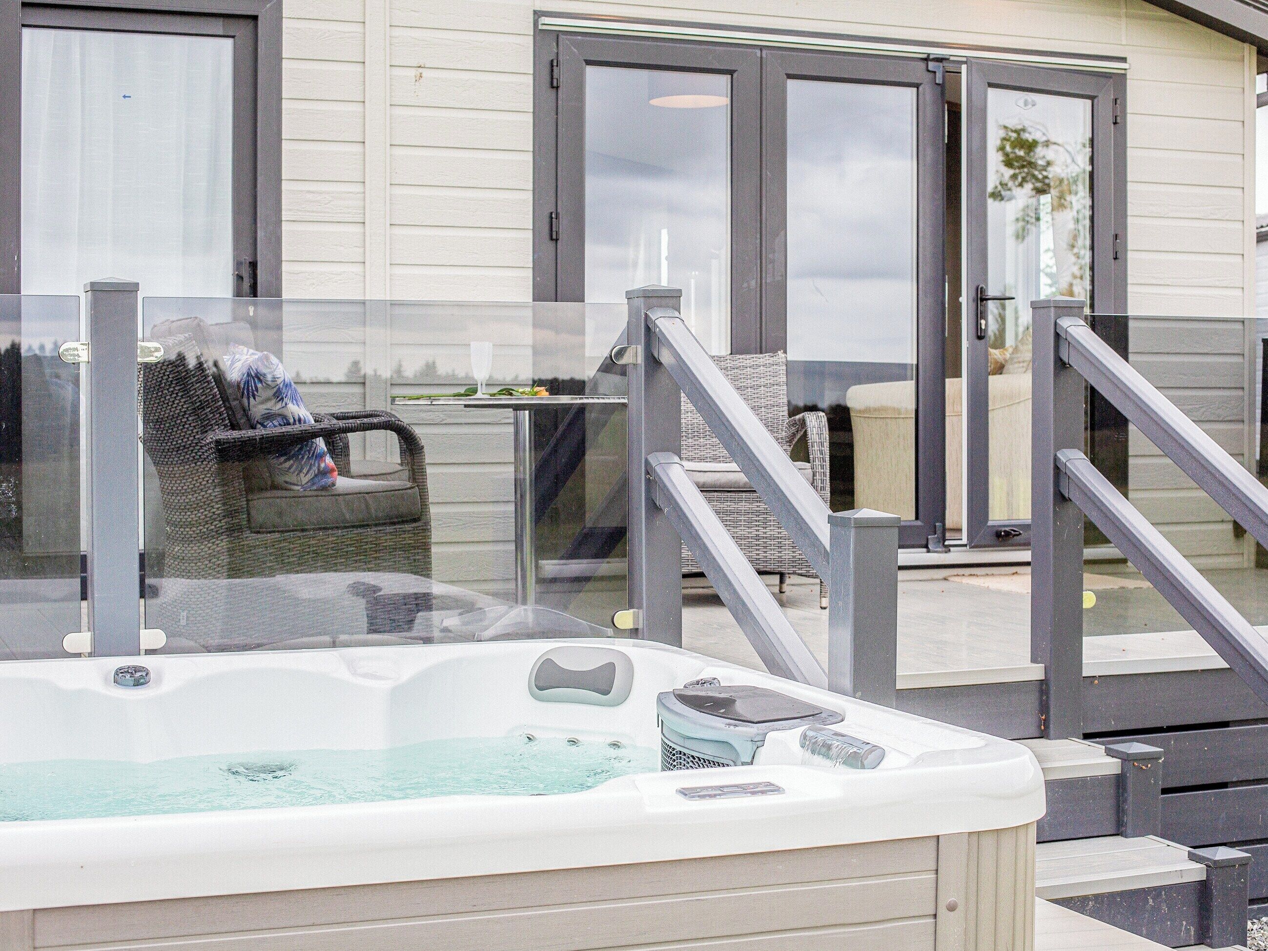 A hot tub with a glass enclosure, outdoor seating area with wicker furniture, and a building with a sliding glass door.