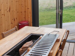 A wooden table with a built-in grill, a red trash bin, and a view of a grassy area through a glass door. - Meikle Balloch - UKC6069 (Newmill, nr Keith)