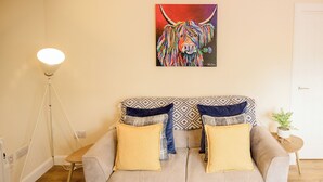 Een woonkamer met een bank, twee kussens, een vloerlamp, een schilderij en een kleine plant.