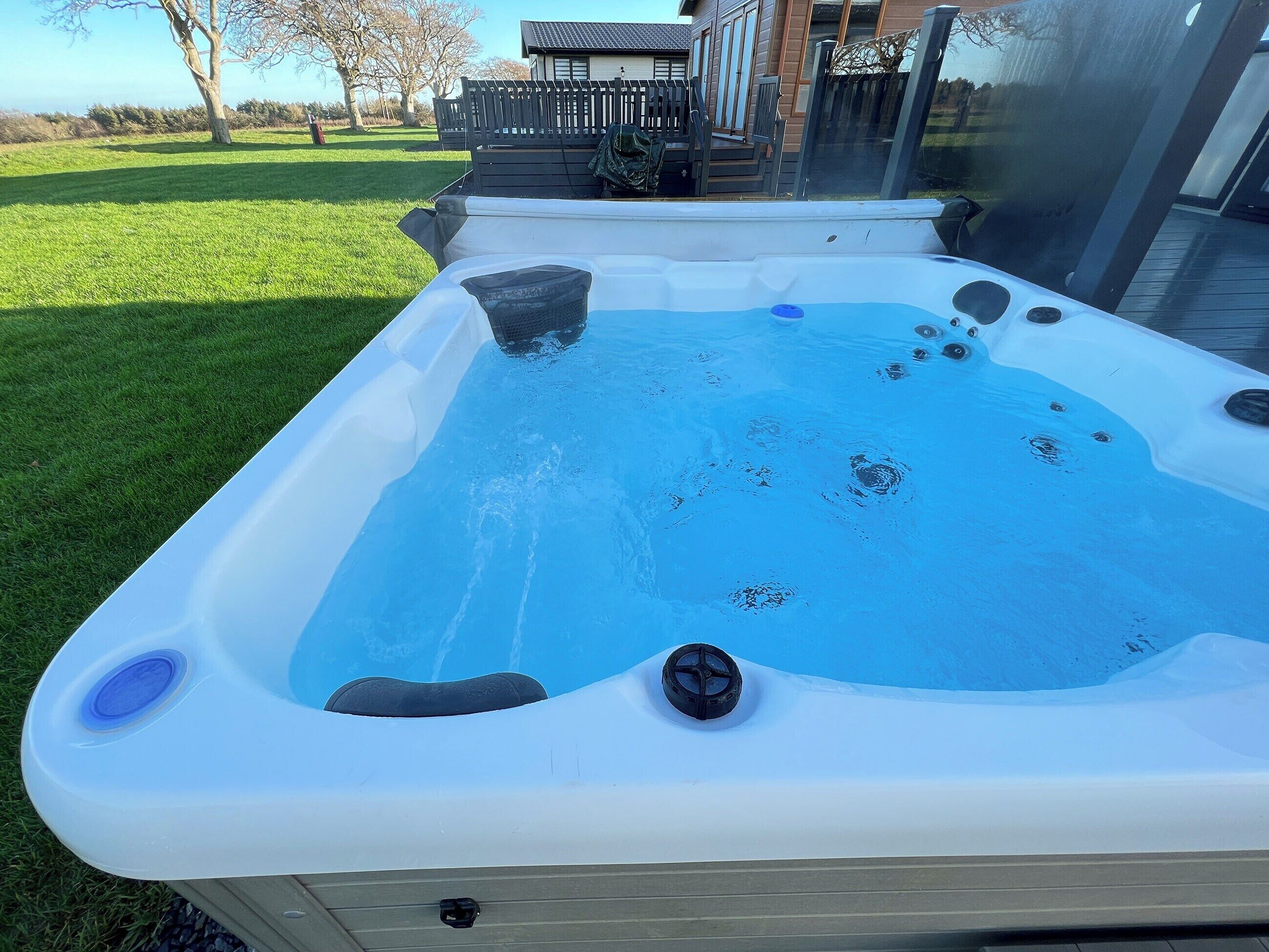 Un jacuzzi con superficie de agua azul cristalina, rodeado por una terraza de madera y un césped.