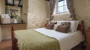 A bedroom with a stone wall, a bed with patterned pillows, a wooden headboard, a bedside table with a lamp, and a window with patterned curtains. - 1 bedroom accommodation in Wooton Fitzpaine, near Charmouth (Wooton Fitzpaine, near Charmouth)