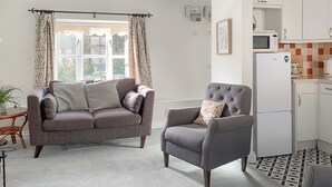 Een woonkamer met een grijze bank, een grijze fauteuil en een klein bijzettafeltje met een plant. Er is een raam met gordijnen, een magnetron en een koelkast in de keuken.
