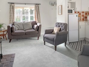 A living room with a grey sofa, a grey armchair, and a small side table with a plant. There is a window with curtains, a microwave, and a refrigerator in the kitchen area. - A wonderful retreat for two couples or a family to enjoy. (Wooton Fitzpaine, near Charmouth)