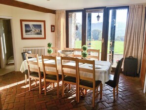 Ein Essbereich mit einem Tisch, gedeckt für sechs Personen, Holzsesseln, einer weißen Tischdecke, einem gerahmten Bild an der Wand und Blick auf eine grüne Landschaft durch die Glastür.