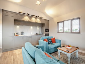 A modern kitchen with grey cabinets, a microwave, and a coffee maker. There is a blue sofa with orange cushions and a small wooden table with fruit. - A water’s edge setting in the heart of the Norfolk Broads National Park. (Brundall)