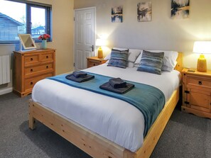 A bedroom with a wooden bed, a nightstand with a lamp, a dresser, and artwork on the wall. - A water’s edge setting in the heart of the Norfolk Broads National Park. (Brundall)